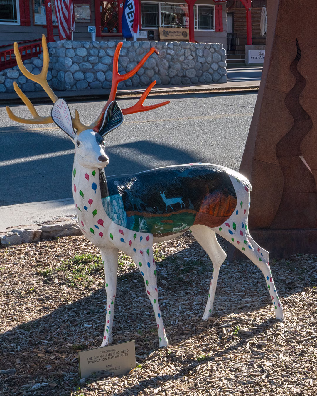 White deer sculpture painted with colorful dots, orange antlers and a mountain scene on its side